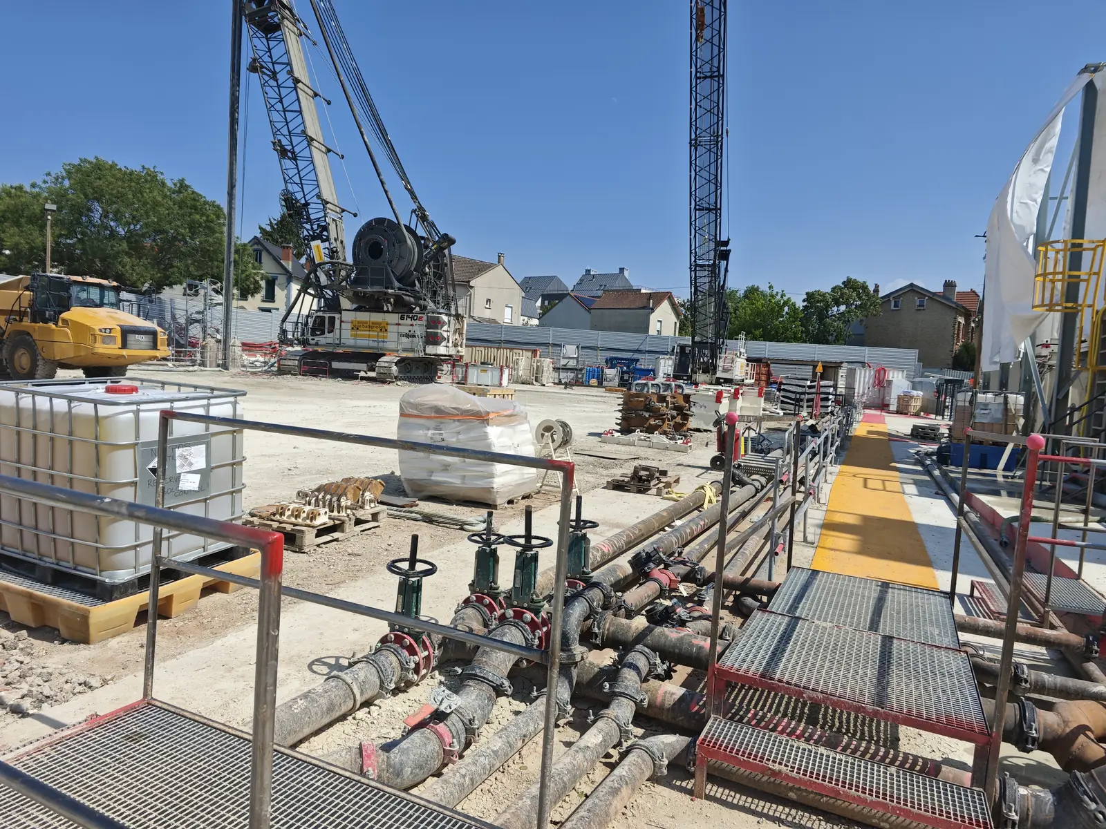 Tuyauteries à vannes au premier plan, passerelles, cuves et engins de chantier en arrière-plan.